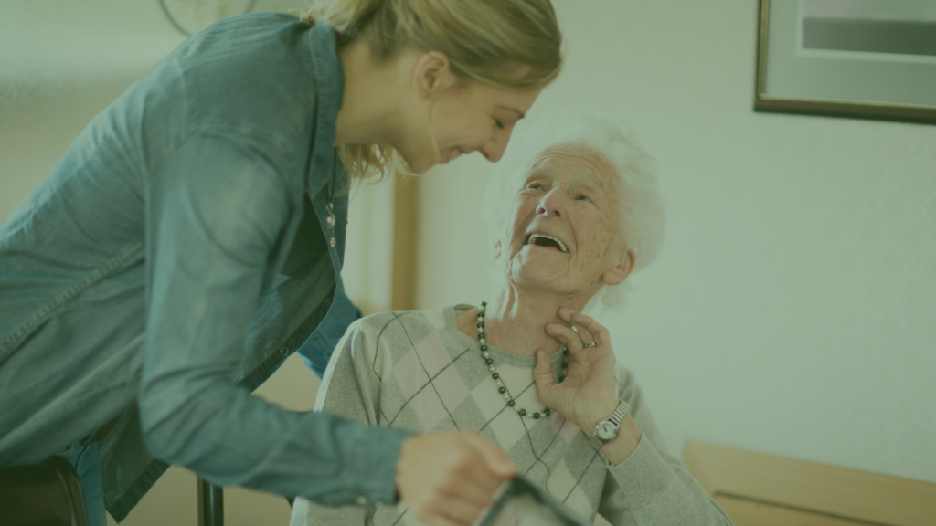 Two women, one is older and in a wheelchair, the other leans over her with a caring smile.