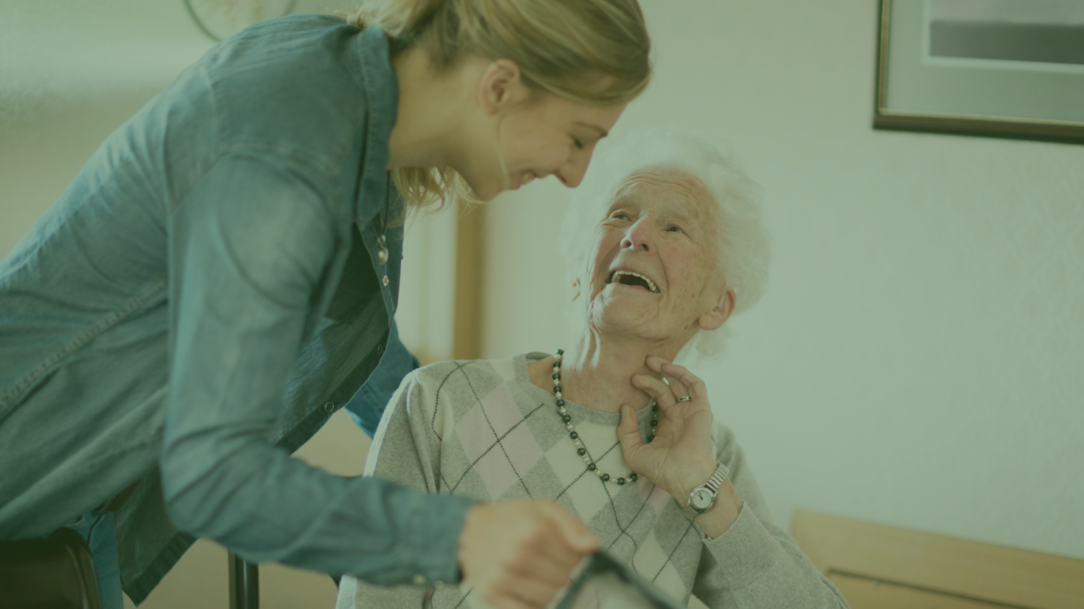 Two women, one is older and in a wheelchair, the other leans over her with a caring smile.