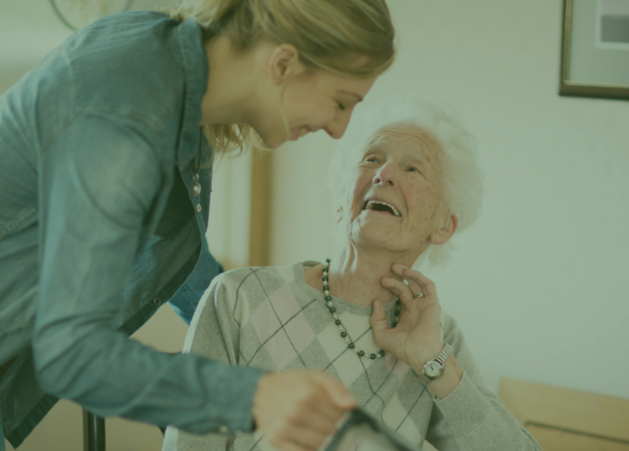 Two women, one is older and in a wheelchair, the other leans over her with a caring smile.