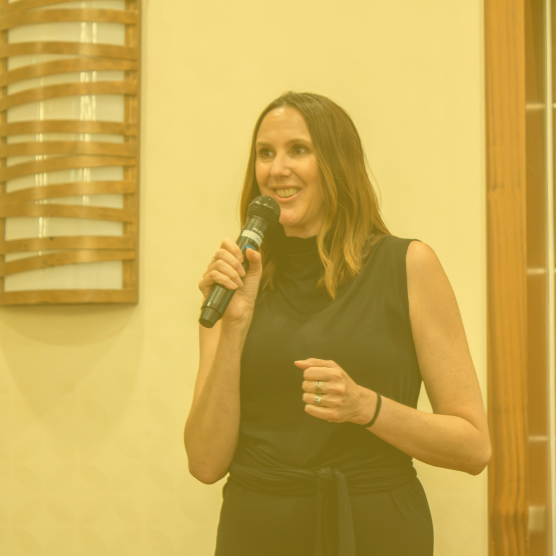 Executive Director, Lori Pfingst speaks into a microphone. Photo has a yellow tinge.